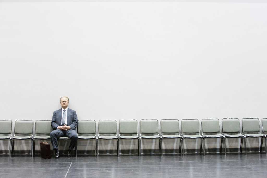 一名身穿蓝色西装的男子在靠着白墙的一排空椅子上等待面试